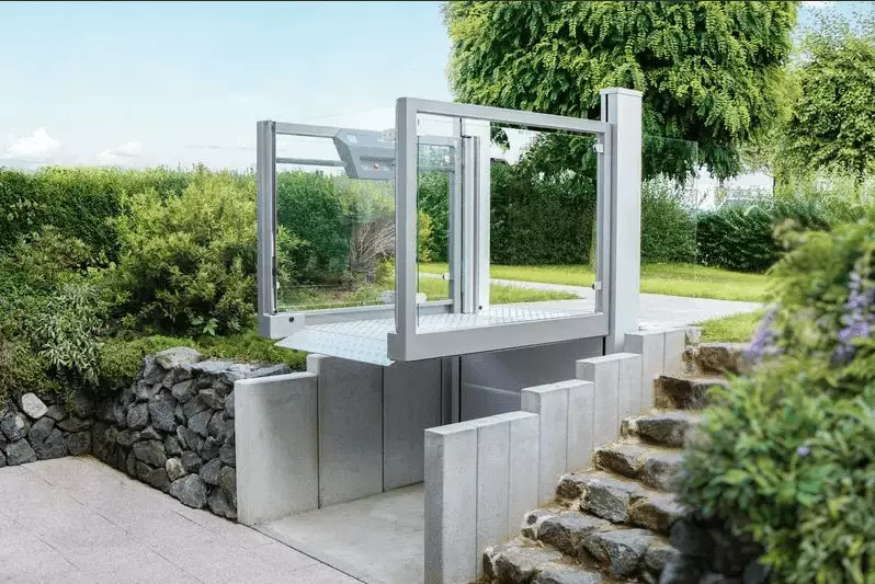 LSM Mobility Plateforme élévatrice verticale Ascendor UnaPorte installée à l'extérieur, reliant une terrasse à un jardin verdoyant via des escaliers. Structure moderne en gris et verre, intégrée dans un aménagement paysager soigné.