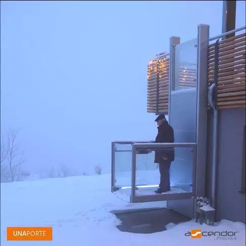 LSM Mobility Un homme utilise la plateforme élévatrice verticale Ascendor UnaPorte dans un paysage enneigé. L'ascenseur extérieur est moderne avec des parois en verre. UNAporte Ascendor Lifttechnik.