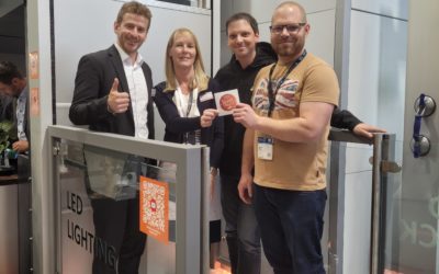 LSM Mobility Quatre personnes souriantes présentent un badge à Interlift à Augsbourg 2022, devant un stand d'éclairage LED. L'ambiance est conviviale et professionnelle.