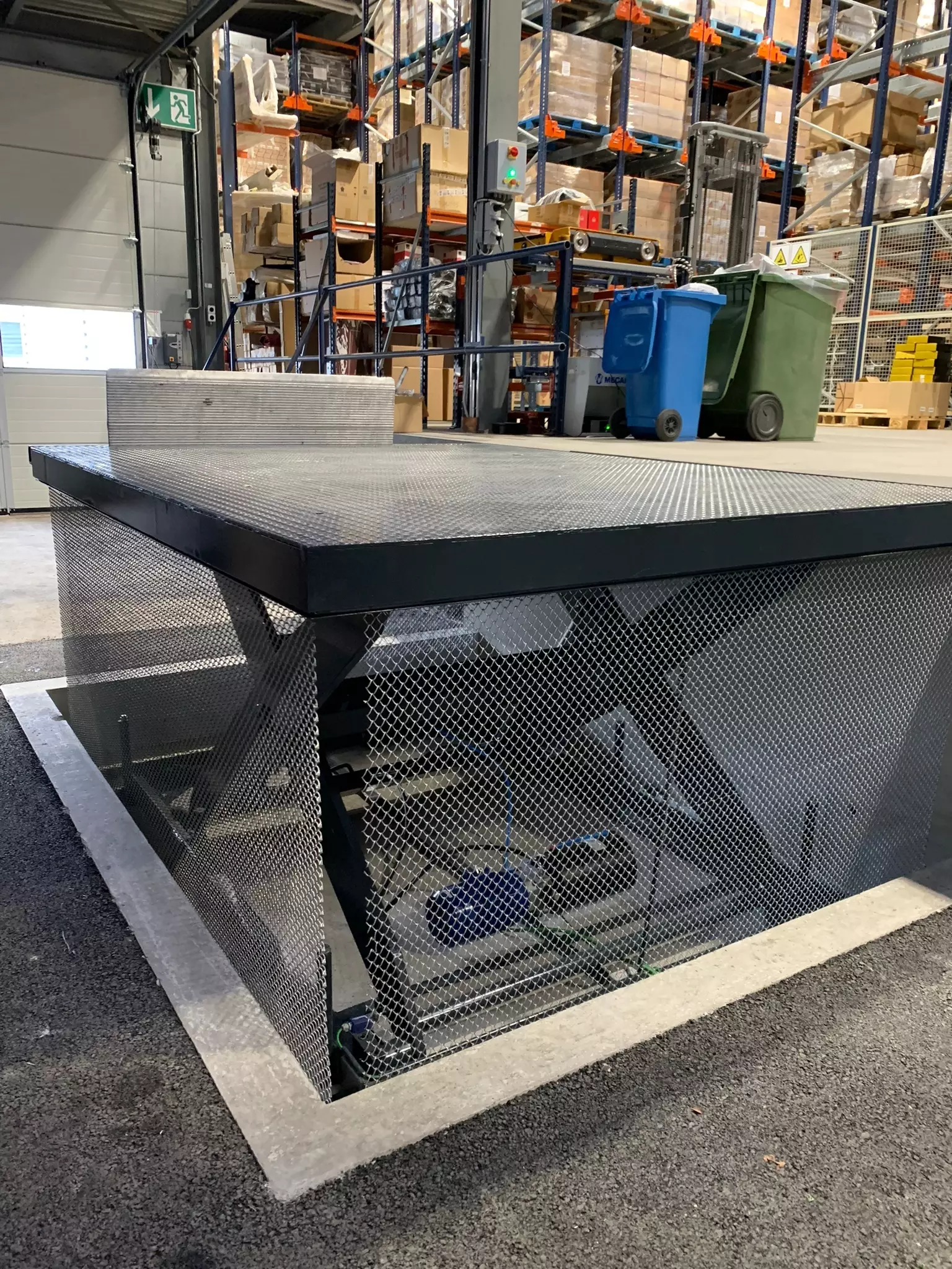LSM Mobility Plateforme industrielle grillagée noire à Puidoux, vue dans un entrepôt avec des rayonnages chargés de cartons. Des poubelles bleues et vertes sont visibles à l'arrière-plan.