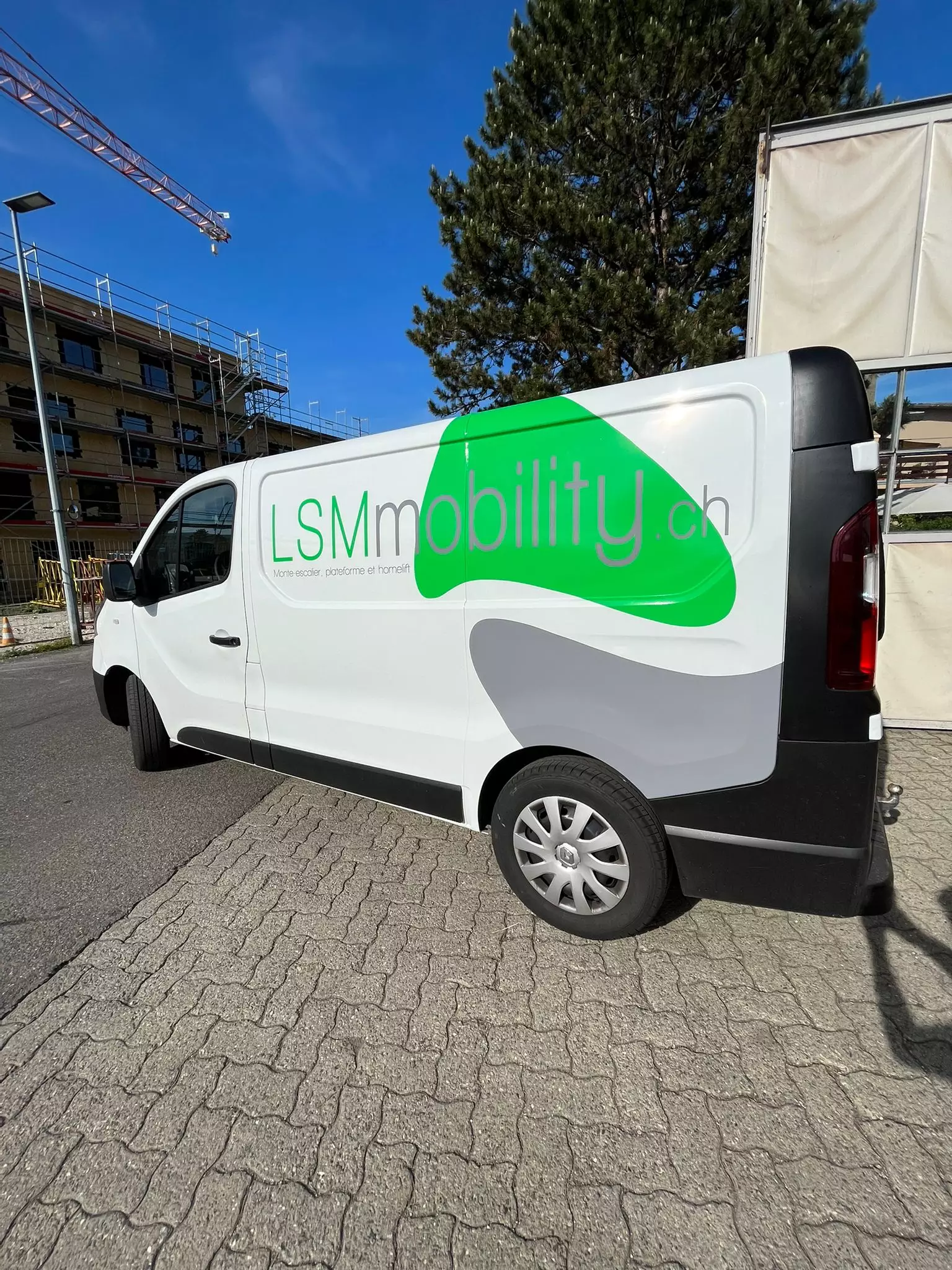 LSM Mobility Nouveau véhicule : Fourgonnette blanche LSMmobility.ch avec logo vert et gris. Bâtiment en construction et ciel bleu en arrière-plan.