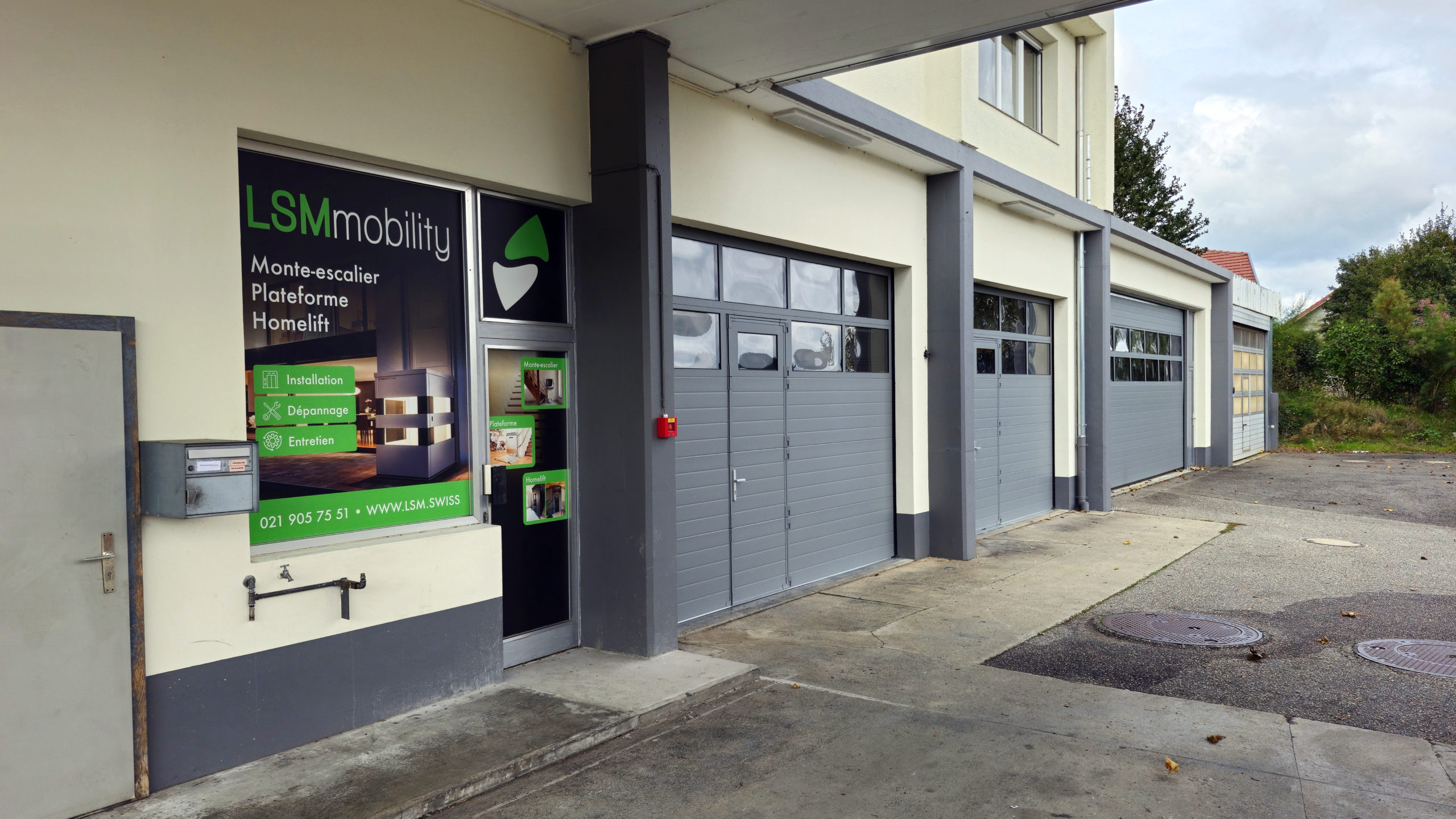 LSM Mobility Façade du nouveau local de LSMobility avec l'enseigne "Monte-escalier Plateforme Homelift". Les portes de garage grises suggèrent un espace de stockage ou d'installation. "Nous avons déménagé!