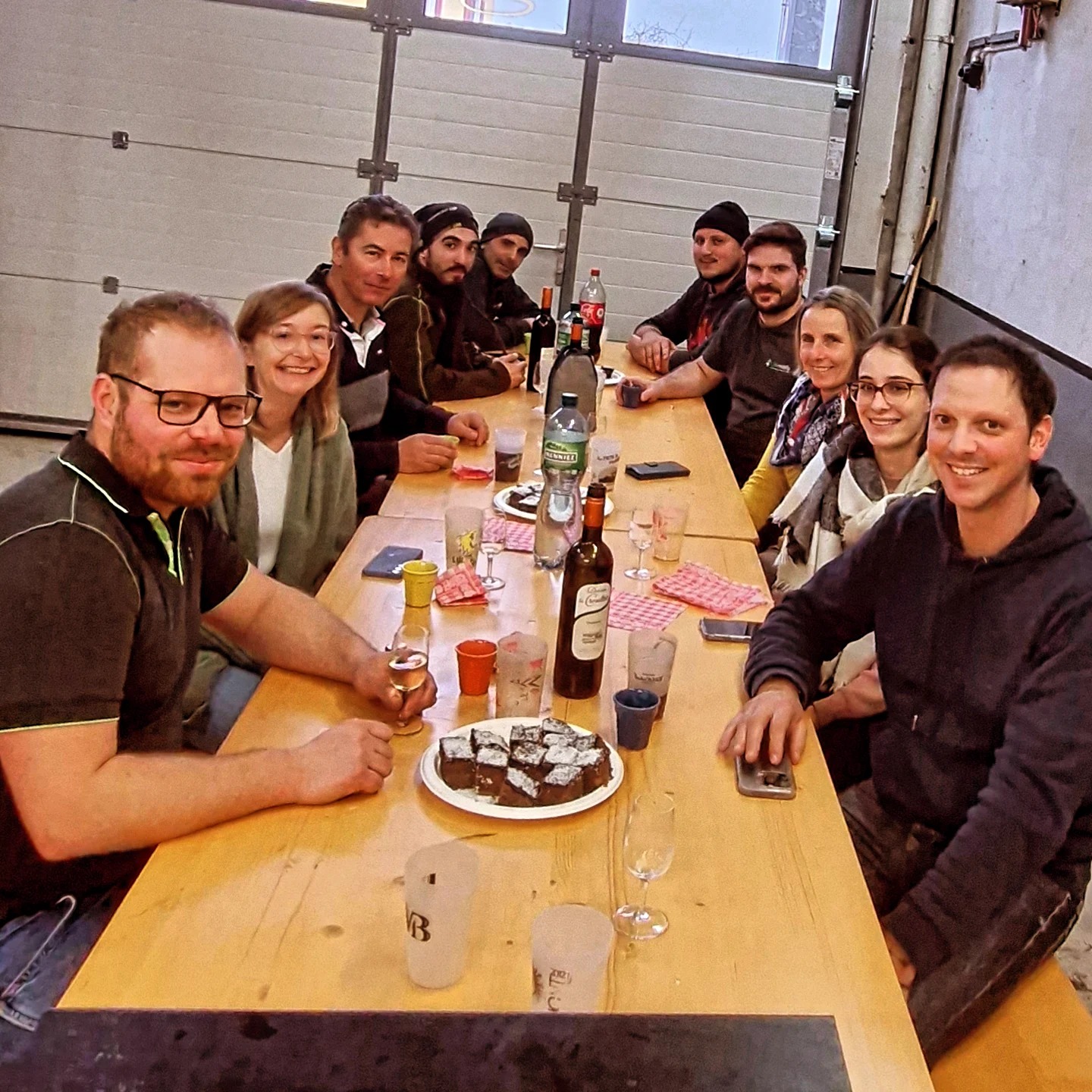 LSM Mobility Un groupe de personnes souriantes assises autour d'une table en bois, partageant une raclette de fin d'année dans une ambiance conviviale. Bouteilles de vin et gâteau au chocolat présents.