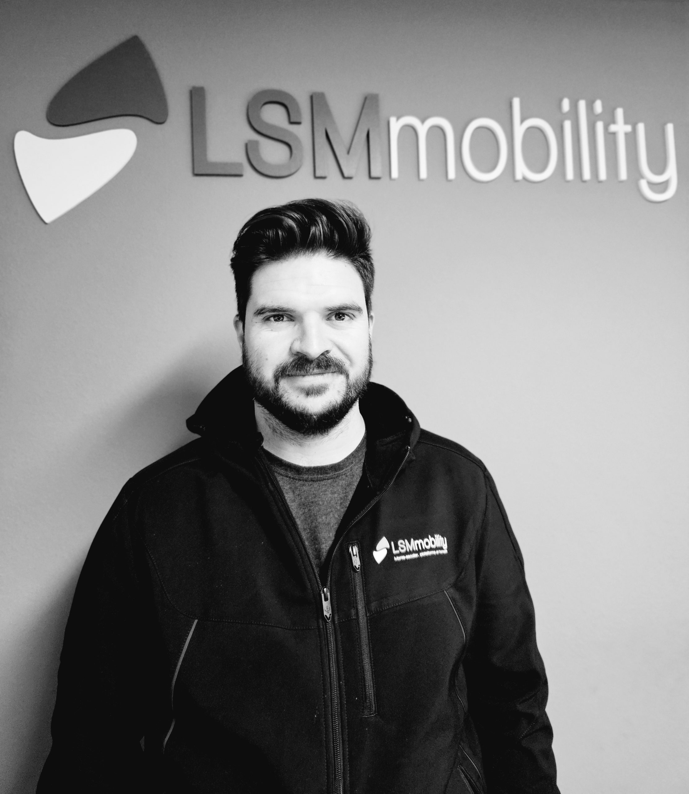 LSM Mobility Un homme souriant, portant une veste LSMmobility, se tient devant le logo de l'entreprise. Présentation d'un nouveau collaborateur.
