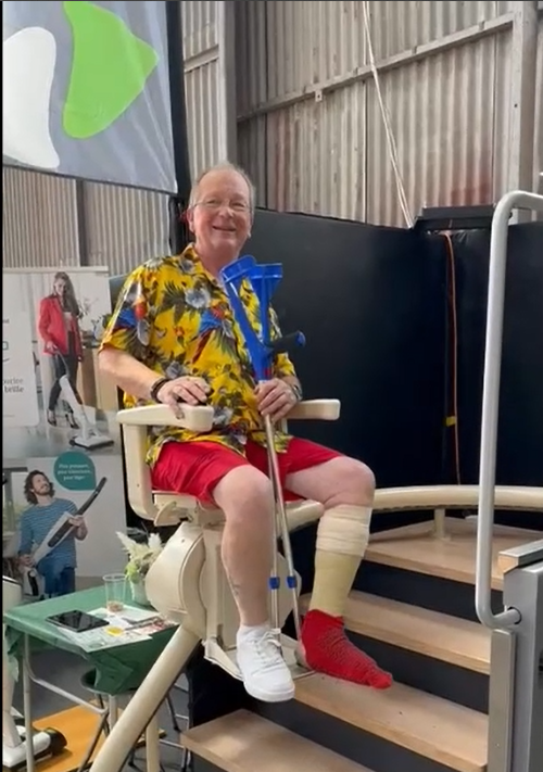LSM Mobility Un homme souriant, en chemise fleurie et short rouge, assis sur un monte-escalier avec des béquilles. Solution pour rester autonome chez soi.